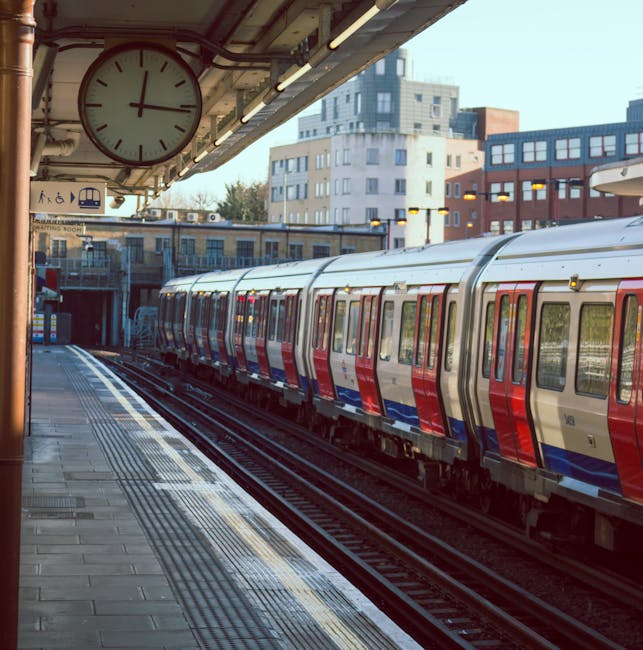 A modern London underground train with a silver exterior featuring red and blue accents is stopped at Elmstead Woods station platform, which is paved with concrete tiles. The platform has tactile paving strips along the edge for safety, and a large round clock displaying the time is mounted under the station’s overhanging roof, which is supported by metal beams. In the background, there are multi-storey residential and commercial buildings made of brick and concrete, with windows reflecting sunlight. Some luggage or boxes may be visible on the platform, and the scene appears to depict a typical day for home relocation or furniture transport, with [COMPANY_NAME] involved in the moving process often associated with packing and loading activities. The lighting suggests daytime, possibly morning or late afternoon, with clear weather and a bright sky overhead. This environment is part of a transportation hub that supports logistics for house removals near Elmstead Woods station, ELMSTEAD.