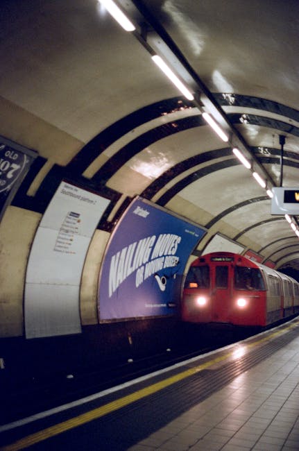 Inside a London Underground station tunnel with curved, tiled walls and overhead lighting, a red train is stationary at the platform. The platform has a tactile paving strip near the edge for safety, and there are large advertisements on the wall behind the train, including one with the text 'MAN WITH A VAN.' The scene suggests a setting related to home relocation or furniture transport, with environmental features typical of modular, underground transport systems. The lighting is bright and the space is clean, emphasizing the station’s modern design, which supports the logistics of moving or transporting household items via the underground network, as would be coordinated by a professional removals service like Man with Van Elmstead.