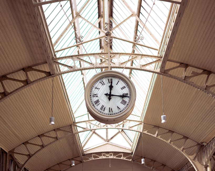 A modern London underground train with a silver exterior featuring red and blue accents is stopped at Elmstead Woods station platform, which is paved with concrete tiles. The platform has tactile paving strips along the edge for safety, and a large round clock displaying the time is mounted under the station’s overhanging roof, which is supported by metal beams. In the background, there are multi-storey residential and commercial buildings made of brick and concrete, with windows reflecting sunlight. Some luggage or boxes may be visible on the platform, and the scene appears to depict a typical day for home relocation or furniture transport, with [COMPANY_NAME] involved in the moving process often associated with packing and loading activities. The lighting suggests daytime, possibly morning or late afternoon, with clear weather and a bright sky overhead. This environment is part of a transportation hub that supports logistics for house removals near Elmstead Woods station, ELMSTEAD.