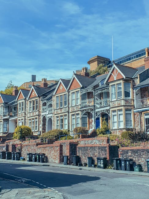 A row of Victorian terraced houses with ornate metalwork balconies and large bay windows located on a street with a brick retaining wall and small front gardens. Several wheelie bins are lined up along the footpath, which leads to the entrances of the properties. The sky above is clear with some wispy clouds, and the scene is bathed in daylight. This residential area illustrates typical housing that might be involved in house removals or home relocation services, as provided by Man with Van Elmstead, situated in Elmstead, with an emphasis on professional furniture transport and packing during the moving process.