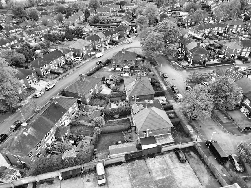 An aerial black-and-white photograph capturing a suburban area with numerous detached and semi-detached houses, most with pitched roofs and gardens. In the foreground, a house has its back garden partially visible, with a driveway and a parked van. Next to the house, a team of movers from Man with Van Elmstead are engaged in a home relocation process, loading cardboard boxes, wrapped furniture, and large household appliances into the back of a large moving van parked on the street outside the property. The van is positioned on the pavement near the entrance of the house, with the movers using hand trucks and blankets to protect furniture during loading. Surrounding the property are paths, trees, and residential streets, with additional vehicles parked nearby. The scene showcases a typical house removal operation focused on furniture transport and packing for a house move, set within a quiet, well-established neighbourhood in Bromley. The detailed environment emphasizes the logistics of moving services, including packing, loading, and transportation, as carried out by Man with Van Elmstead.