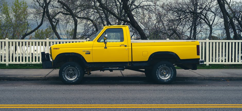 A yellow pickup truck parked on the side of a suburban street in front of a white wooden fence, with tall, leafless trees in the background during daytime. The truck's bed is empty, and the vehicle appears to be used for home relocation or furniture transport services by Man with Van Elmstead, as part of their removals and moving logistics. The street has a double yellow line near the curb, and the scene is well-lit with natural daylight, illustrating the vehicle ready for loading or unloading furniture and boxes as part of packing and moving processes.