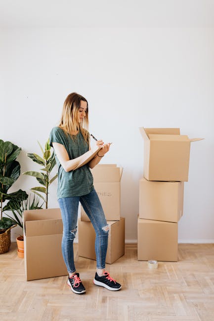A woman with shoulder-length blonde hair, dressed in a casual grey t-shirt and ripped jeans, stands inside a room next to several stacked cardboard moving boxes, some open and others closed. She is holding a pen and a small notepad, appearing to be taking notes or checking inventory amid her home relocation process. The room has a light-colored wooden floor and plain white walls, with a large potted plant situated nearby on the left side of the image. A small orange pot is visible on the floor next to the plant. The scene suggests the packing and moving process, with the woman engaged in organizing or planning her move, supported by the presence of packaging materials and boxes ready for transport. The environment is well-lit, emphasizing the practical aspects of packing furniture, household items, or personal belongings as part of a professional removal service by Man with Van Elmstead.