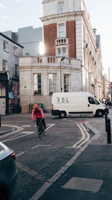 A cyclist wearing a red helmet and pink jacket rides a bicycle across a narrow street in Elmstead. In the background, a white van with the initials 'RSL' is parked adjacent to a multi-storey residential property built with red brick and cream stone detailing, characteristic of London architecture. The van is positioned near the entrance of a property, possibly being loaded or unloaded during a home relocation. The scene is lit by natural daylight, casting shadows on the pavement, with nearby buildings including a mix of modern and traditional structures. This image illustrates aspects of urban moving logistics, such as transporting furniture or boxes, with the van possibly used by Man with Van Elmstead for removals. The street features white and yellow road markings, and a bicycle lane is visible alongside the vehicle and building facades, capturing typical elements involved in residential relocations in a city environment.