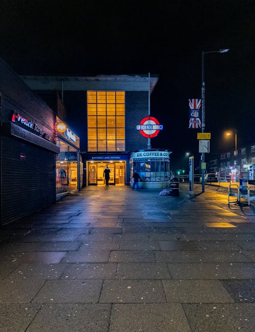 A nighttime scene outside a train station with illuminated signs reading 'Underground' and 'Tube,' and a bright orange-lit window above the entrance. The station is located on a narrow street with a pavement made of rectangular stone tiles, reflecting wetness from recent rain. To the right, there are shop fronts with closed shutters, and a streetlamp casting yellow light onto the pavement. A small group of people are visible near the station entrance, some standing and some walking, with their figures slightly blurred. On the right side, a bus stop shelter and a vertical street sign are also visible. The overall environment suggests a busy urban area suitable for house removals or furniture transport, with well-lit transport links and a typical street layout. The scene captures a stable, secure setting where professional movers, such as [COMPANY_NAME], might operate during evening home relocation or moving services in this area.