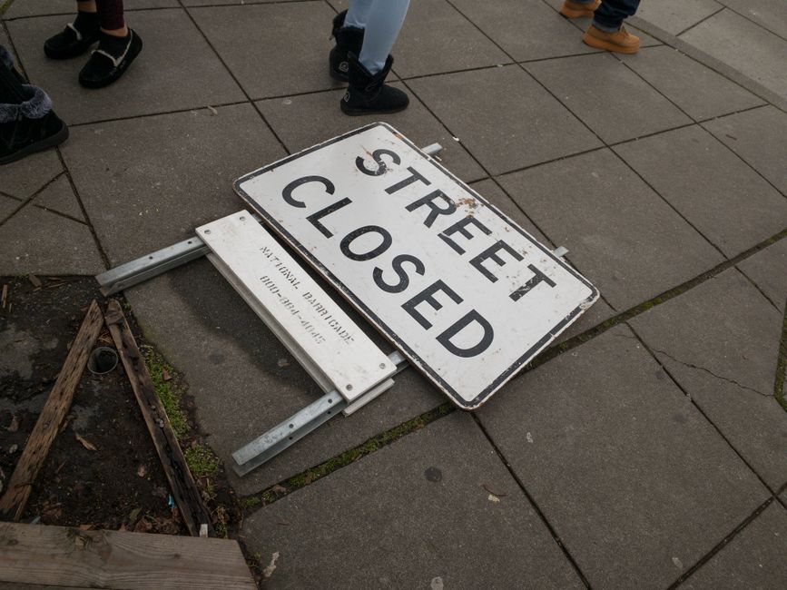 A fallen rectangular metal street closure sign with a white background and black lettering reading 'STREET CLOSED' lies on a paved sidewalk next to a small dirt area with wooden edging, which contains some soil and a few small plants. The sign is attached to a metal frame with two supports, one of which is partially visible. Several people's footwear, including sneakers and boots, are seen standing nearby on the pavement. The scene appears to be in an urban area with tiled pavement, and the fallen sign indicates temporary street closure, possibly related to local regulations or work permit enforcement, as suggested by the webpage about street-closure rules and potential fines. The image is relevant to house removals and moving logistics, illustrating the importance of coordination and awareness of street restrictions during home relocation activities, with [COMPANY_NAME], Man with Van Elmstead, operating within the context of removals.