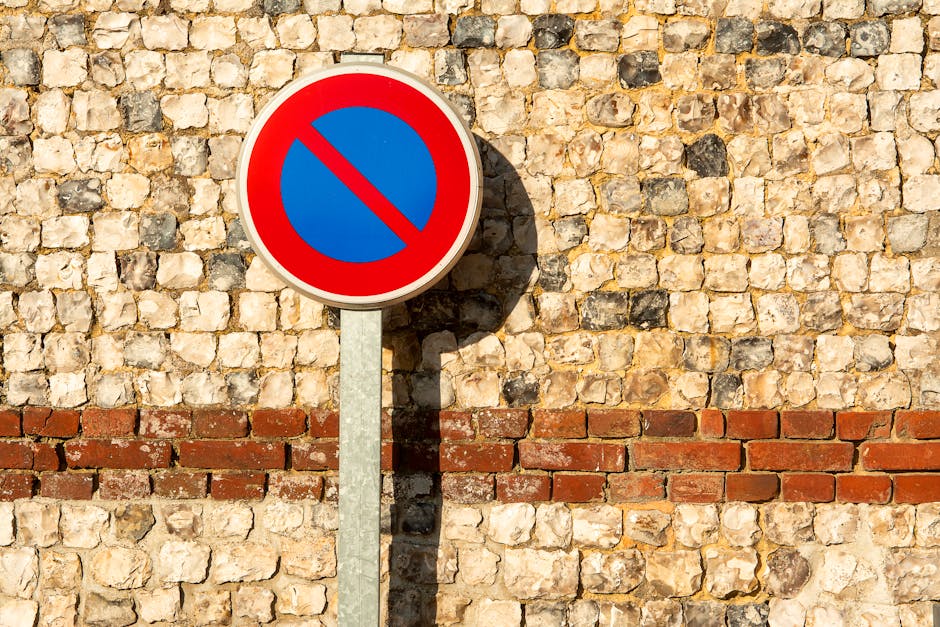 A round no parking traffic sign with a red border and a blue background featuring a red diagonal line, mounted on a metal pole casting a shadow on a textured brick wall made of beige and light brown stones. The wall has a horizontal strip of red bricks running across its middle. The sign is positioned on the pavement outside a residential property, indicating parking restrictions that may impact home relocation or furniture transport processes. This scenario relates to house removals and moving logistics, where parking regulations can influence loading and unloading activities. The image, associated with Man with Van Elmstead, exemplifies the types of signage that may be encountered during local moves or relocation services, highlighting the importance of understanding street rules and potential fines during the moving process.
