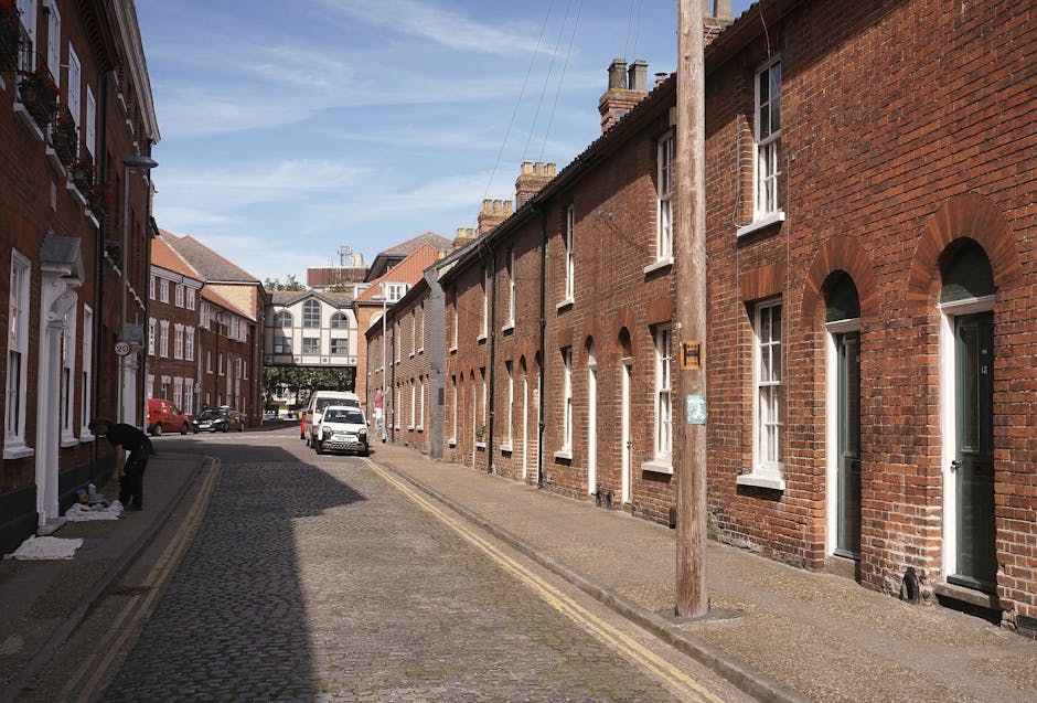 A narrow cobbled street in Elmstead with rows of red brick terraced houses on both sides, each featuring white-framed windows and front doors. On the left, a person is seen packing or unpacking items on a mat outside a house. Several cardboard boxes and packing materials are visible, suggesting an ongoing move. A white van is parked on the street, with its back doors open, likely being loaded or unloaded as part of a home relocation or furniture transport process. Utility poles and street lamps line the sidewalk, and in the background, a modern building with large glass windows spans across the street, connecting the two sides. The scene is lit by natural daylight, with shadows cast on the street, reflecting typical packing and moving activities facilitated by companies like Man with Van Elmstead, involved in residential removals and relocation services.