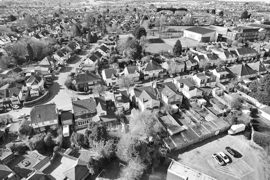 An aerial black-and-white photograph showing a residential neighbourhood with multiple detached and semi-detached houses arranged along winding streets. The houses vary in size and style, some with visible chimneys, gabled roofs, and front gardens. Several trees and greenery are interspersed among the homes, with some trees casting shadows on the properties. A large open sports field or park with a sports hall is visible in the background. In the foreground, a driveway leads to a parking area where a few vehicles are parked, including a van likely used for home relocation. The scene captures the process of house removals and furniture transport, with some houses surrounded by packing materials like plastic wraps and cardboard boxes, indicating ongoing or planned moving activities. The photo is a detailed view of a typical suburban area, focusing on the layout of houses, vehicle positioning for moving logistics, and the surrounding environment, all within a context that Man with Van Elmstead might service for end-of-tenancy removals.
