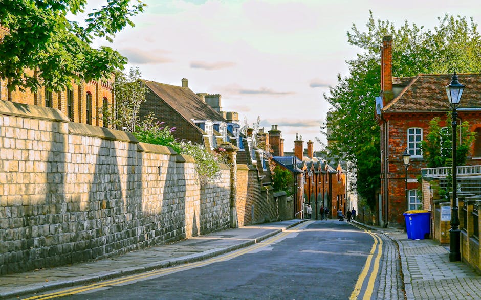 A narrow cobbled street in Elmstead with rows of red brick terraced houses on both sides, each featuring white-framed windows and front doors. On the left, a person is seen packing or unpacking items on a mat outside a house. Several cardboard boxes and packing materials are visible, suggesting an ongoing move. A white van is parked on the street, with its back doors open, likely being loaded or unloaded as part of a home relocation or furniture transport process. Utility poles and street lamps line the sidewalk, and in the background, a modern building with large glass windows spans across the street, connecting the two sides. The scene is lit by natural daylight, with shadows cast on the street, reflecting typical packing and moving activities facilitated by companies like Man with Van Elmstead, involved in residential removals and relocation services.