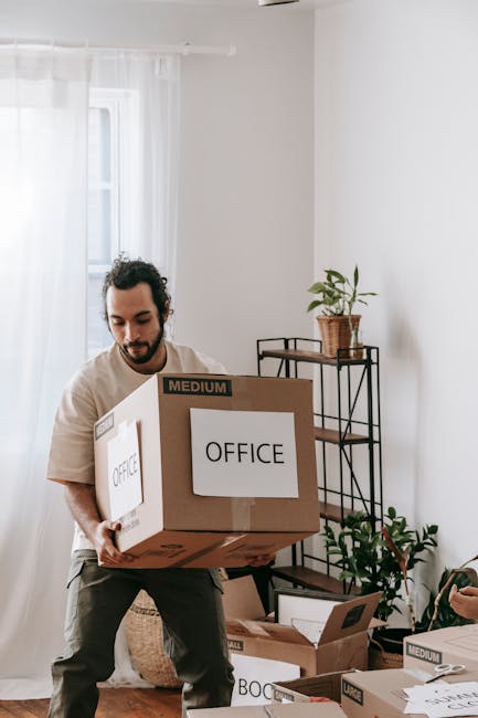 A man moving office supplies inside a bright, minimalist room with white walls and sheer curtains covering a window. He is carrying a medium-sized cardboard box labeled 'OFFICE' and wearing casual clo