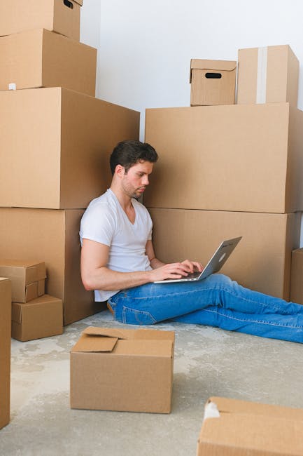 A man moving office supplies inside a bright, minimalist room with white walls and sheer curtains covering a window. He is carrying a medium-sized cardboard box labeled 'OFFICE' and wearing casual clo